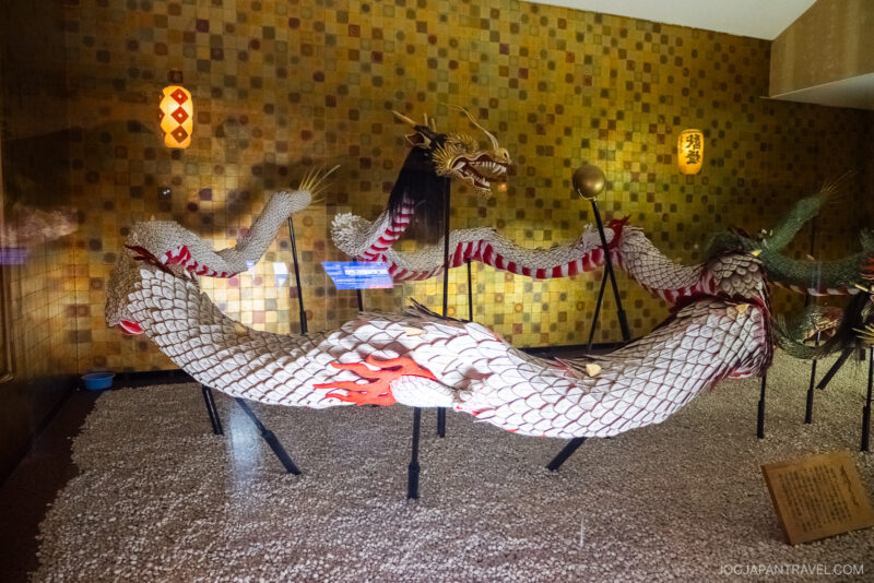 A large, white dragon sculpture with red markings and gold accents is displayed on stands in a room at Glover Garden, featuring a yellow-tiled wall and two lanterns. Its long body coils elegantly over a pebble-covered floor.