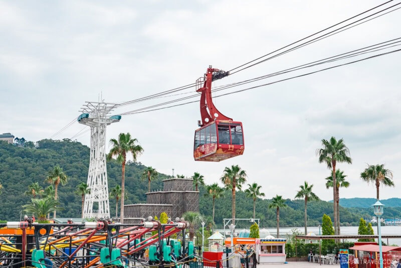 A red cable car travels along suspended cables above Hamanako Pal Pal amusement park, with palm trees, colorful rides, and a distant view of green, forested hills and water.