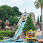 Hamanako Pal Pal (浜名湖パルパル) – Family Fun by Lake Hamana A water ride boat descends a steep blue track on a rocky hill surrounded by greenery at Hamanako Pal Pal amusement park. The Wild River sign is visible in the foreground near palm trees and other park structures.