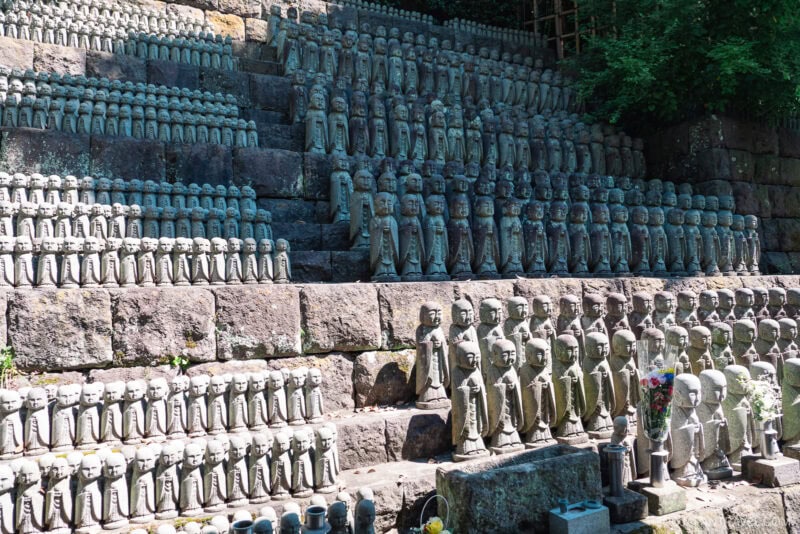 Rows of small stone Jizo statues are arranged on stone steps outdoors at Hasedera, with green foliage in the background. The statues, grouped closely together, vary slightly in size and detail.