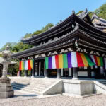 Hasedera Temple (長谷寺) in Kamakura – Temple of Flowers and Mercy A traditional Japanese temple like Hasedera, with ornate, tiered roofs, is decorated with colorful hanging banners. A stone lantern stands in front, and the scene is bright and sunny with lush greenery in the background.