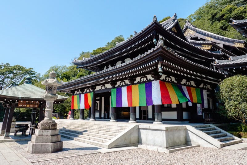 A traditional Japanese temple like Hasedera, with ornate, tiered roofs, is decorated with colorful hanging banners. A stone lantern stands in front, and the scene is bright and sunny with lush greenery in the background.