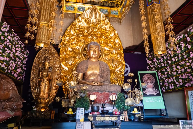 A large golden Buddha statue sits on an ornate pedestal in Hasedera Temple, surrounded by intricate gold decorations, floral arrangements, and offerings. Nearby, a smaller gold statue and informational signs are displayed.