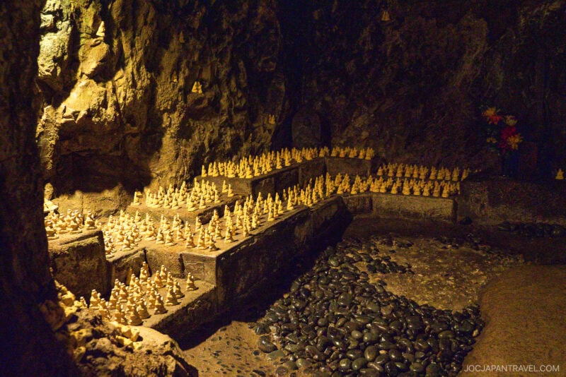 Small golden Buddha statuettes line stone shelves inside a dimly lit cave with rocky walls at Hasedera; black pebbles cover part of the cave floor and artificial flowers are placed in the background.