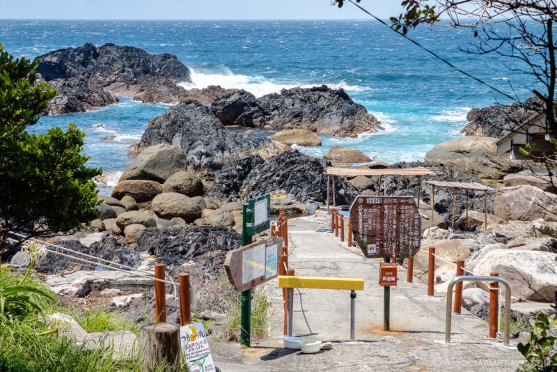 A concrete pathway with signs leads through rocks toward the seaside near Hirauchi Kaichu Natural Onsen, where waves crash against dark, jagged rocks under a bright blue sky. Vibrant turquoise water and lush greenery frame the scenic walkway.