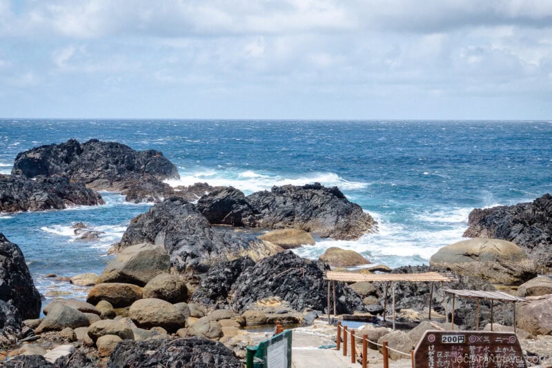 Rocky coastline with waves crashing against dark rocks under a partly cloudy sky. In the foreground, a wooden walkway and sign lead toward Hirauchi Kaichu Natural Onsen, with the ocean stretching out in the background.
