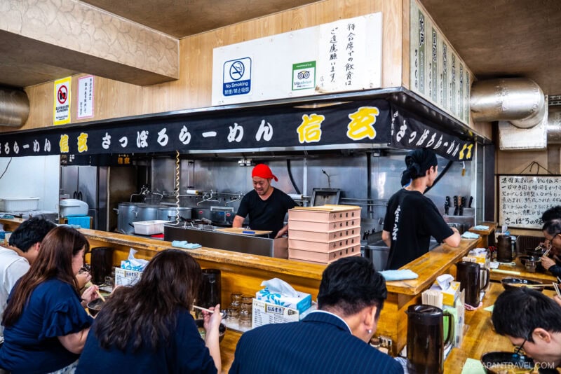 A busy ramen shop in Japan with customers seated at a wooden counter enjoying Sapporo Ramen, while staff prepare food behind the counter. Japanese signs and menus are visible above the kitchen area.