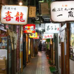 Hokkaido Ramen – Sapporo’s Signature Comfort Food A narrow alleyway in Japan lined with small restaurants and bars, some serving authentic Sapporo Ramen, is illuminated by colorful, glowing Japanese signs and lanterns at night. The passage appears inviting and lively.