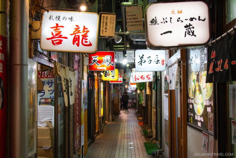 A narrow alleyway in Japan lined with small restaurants and bars, some serving authentic Sapporo Ramen, is illuminated by colorful, glowing Japanese signs and lanterns at night. The passage appears inviting and lively.