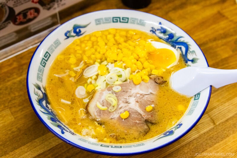 A bowl of Sapporo Ramen with corn, sliced green onions, a piece of pork, and a soft-boiled egg in a rich broth, served in a decorative blue and white bowl with a white spoon.