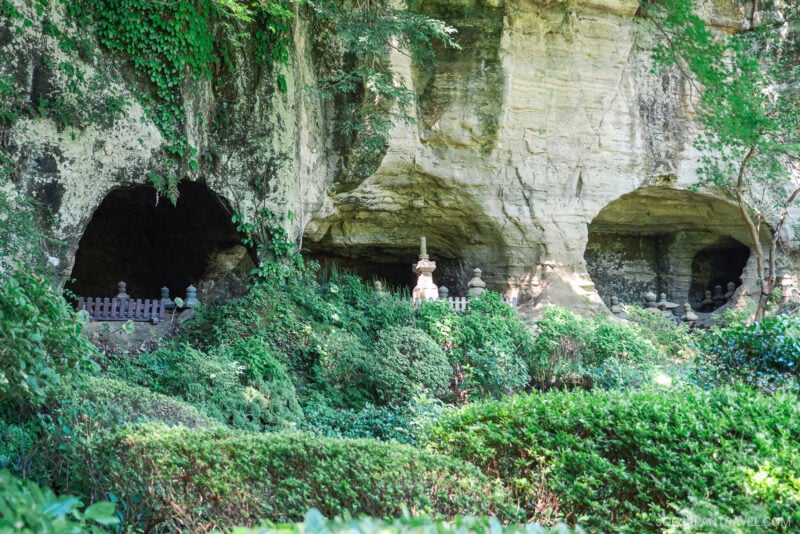 Three cave entrances in a rocky cliff at Hokokuji Temple are surrounded by lush green foliage and bushes, with small stone lanterns and fence-like structures in front of the caves.