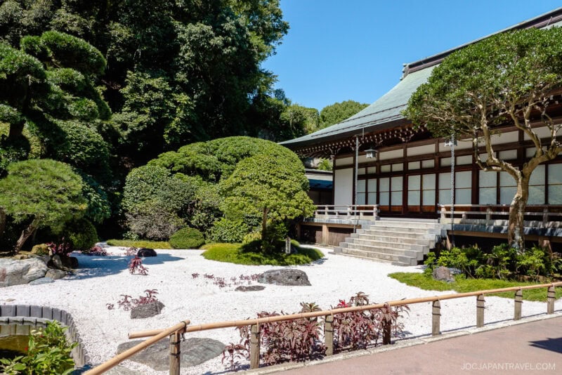 A traditional Japanese zen garden with raked white gravel, manicured bushes, and rocks beside a wooden building with sliding doors, surrounded by greenery at Hokokuji Temple.