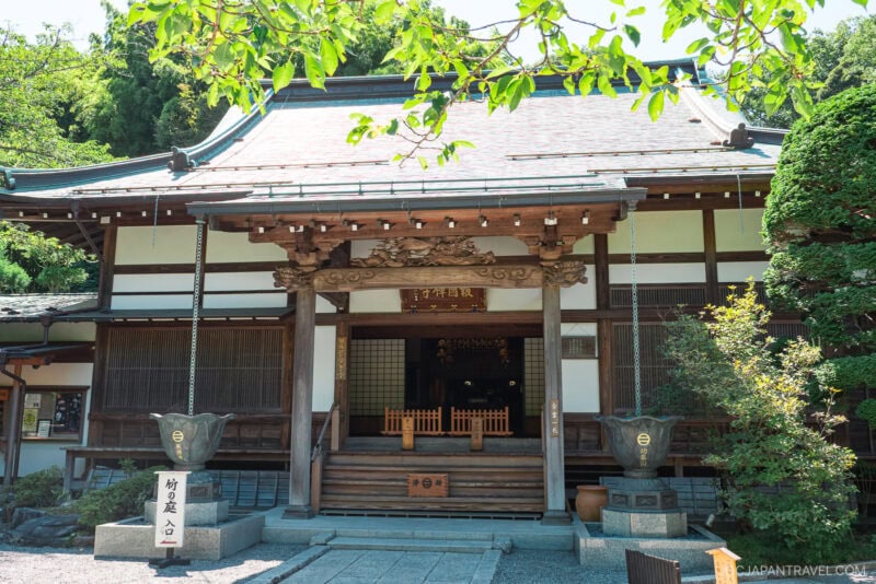 The traditional Japanese wooden Hokokuji Temple, adorned with ornate carvings, stands surrounded by greenery, with stone lanterns and a shaded entrance pathway beneath leafy branches.