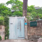 Hollander Slope (オランダ坂 ) – A Stroll Through Nagasaki’s Foreign Settlement Past A pale blue wooden gate set in a stone wall on Hollander Slope, surrounded by lush greenery and trees, with a plaque and sign on the wall; a narrow stone path leads up on the left side.