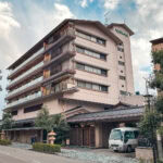 Hotel Tachibana Shikitei (たちばな四季亭) – A Refined Stay in Yamashiro Onsen Hotel Tachibana Shikitei, a multi-story traditional Japanese ryokan with wooden balconies and a tiled roof, stands beside a quiet street with trees and a white van parked at the entrance under a partly cloudy sky.