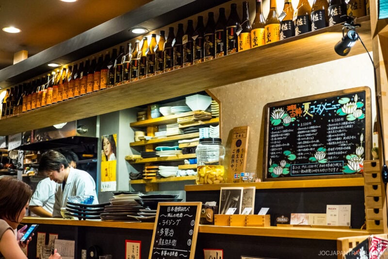 A Japanese restaurant interior with shelves of sake bottles, stacked plates, a handwritten menu board, and a chef working behind the counter&mdash;warm lighting creates a cozy atmosphere, making it a must-visit spot on any Where to Eat in Okayama list.