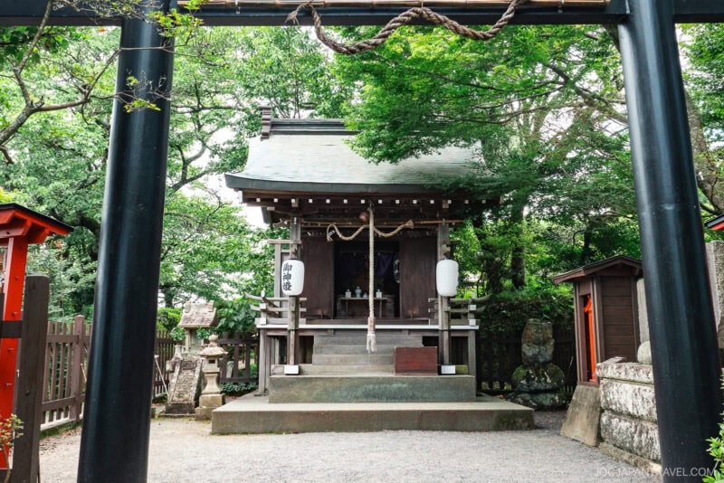 A small Shinto shrine surrounded by trees, with a black torii gate in the foreground and traditional stone lanterns—nestled near Izunokuni Panorama Park, this tranquil spot is framed by classic wooden structures.