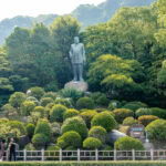 Uncovering the Legacy of Saigo Takamori (西郷隆盛) – Kagoshima’s Last Samurai A large statue of Saigo Takamori stands on a stone pedestal surrounded by manicured bushes and lush green trees, with a mountain in the background and a few people walking in the foreground.