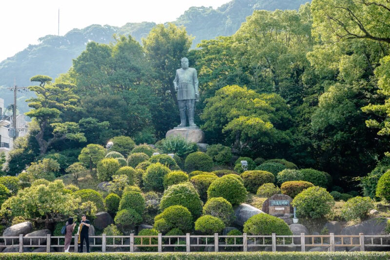 A large statue of Saigo Takamori stands on a stone pedestal surrounded by manicured bushes and lush green trees, with a mountain in the background and a few people walking in the foreground.