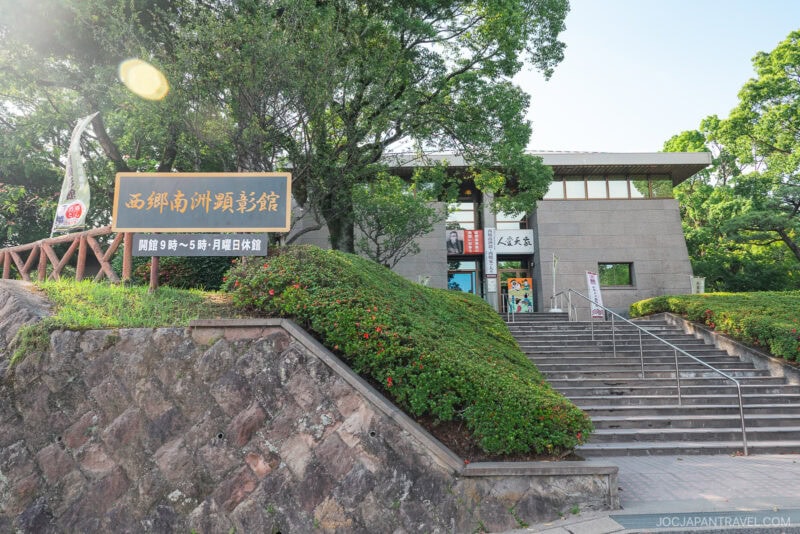 A stone building with large windows sits behind a staircase lined with greenery. Near the entrance, signs with Japanese text reference Saigo Takamori, while sunlight filters through the trees surrounding the scene.