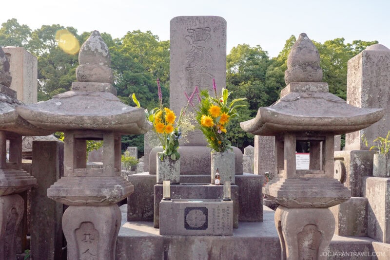 A traditional Japanese grave, said to honor Saigo Takamori, features stone lanterns on either side, fresh yellow flowers in vases, and tall gravestones in the background, set outdoors with trees behind.