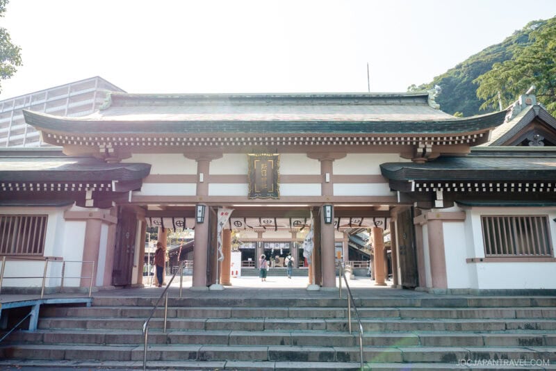 A traditional Japanese shrine entrance with wooden pillars and a tiled roof at Kagoshima Terukuni Shrine, featuring a gold sign above the main gate and steps leading up to the entrance. Two people are visible inside the gate.