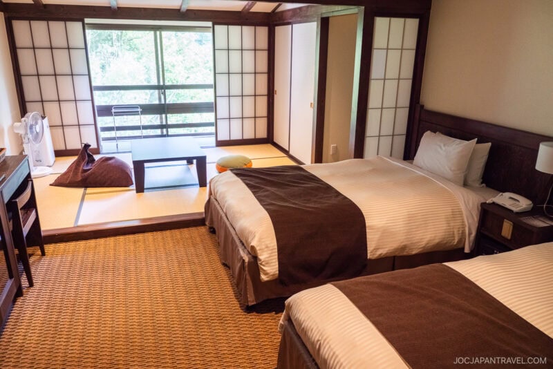 A Kamikochi Alpen Hotel room with two twin beds, shoji sliding doors, a tatami mat area featuring a low table and cushions, a fan, and a large window overlooking lush greenery.