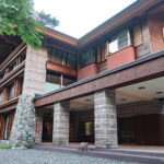 A large, modern building with stone and wood exterior, wide windows, and a sheltered entrance. Surrounded by trees and greenery, the Kamikochi Alpen Hotel features a dog standing near the entrance on a gravel path.