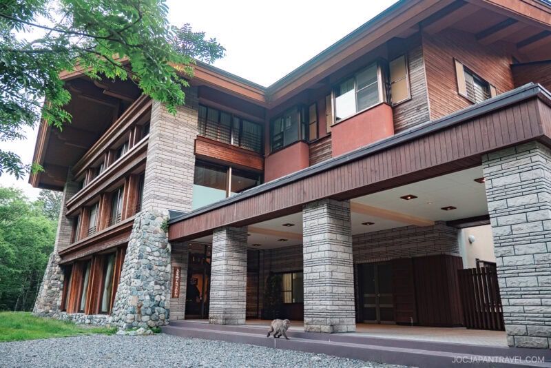 A large, modern building with stone and wood exterior, wide windows, and a sheltered entrance. Surrounded by trees and greenery, the Kamikochi Alpen Hotel features a dog standing near the entrance on a gravel path.