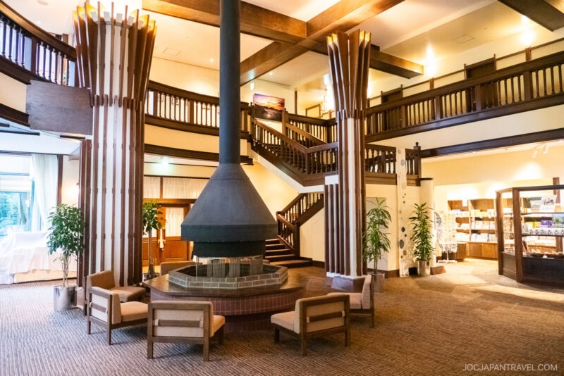 Spacious lobby at Kamikochi Alpen Hotel with a central hanging fireplace, wooden beams, staircase, modern chairs, tall plants, and a gift shop area in the background. Natural light pours in through large windows.