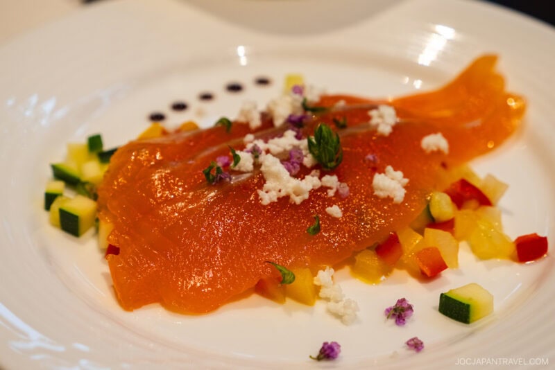 A plate of smoked salmon garnished with crumbled cheese, chopped herbs, edible flowers, and diced colorful vegetables, arranged artfully on a white dish at Kamikochi Alpen Hotel.