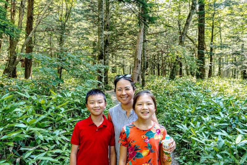 A woman and two children stand smiling on a lush forest trail near Hotaka Shrine, surrounded by green foliage and tall trees in bright daylight. The boy wears a red shirt, and the girl wears a colorful floral shirt.