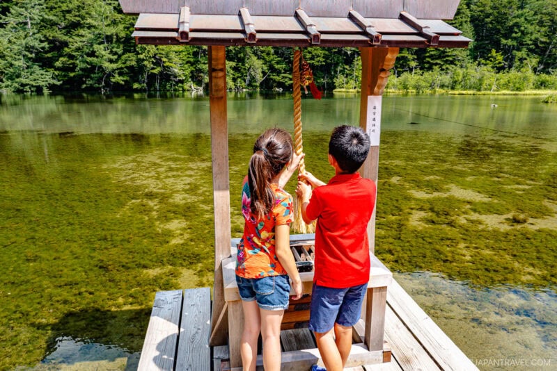 Two children stand on a wooden platform by a clear lake, ringing a bell at the small Hotaka Shrine with a thatched roof, surrounded by lush green trees on a sunny day.