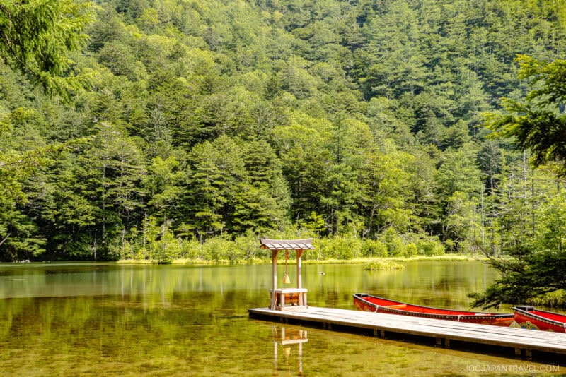 A wooden dock with a small shelter extends into a calm, clear lake near Hotaka Shrine, surrounded by dense green forest; two red canoes are tied to the dock, and the scene is bright and peaceful.
