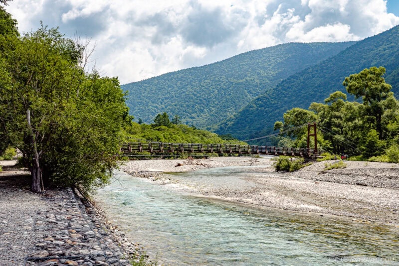 A wooden footbridge crosses a shallow, clear river surrounded by trees, with green mountains—including the area near Hotaka Shrine—rising in the background under a partly cloudy sky.