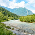 Kamikochi – Hike to Taisho Pond (大正池) A clear river flows through lush green forests, with tall, tree-covered mountains and Taisho Pond in the background under a partly cloudy sky.