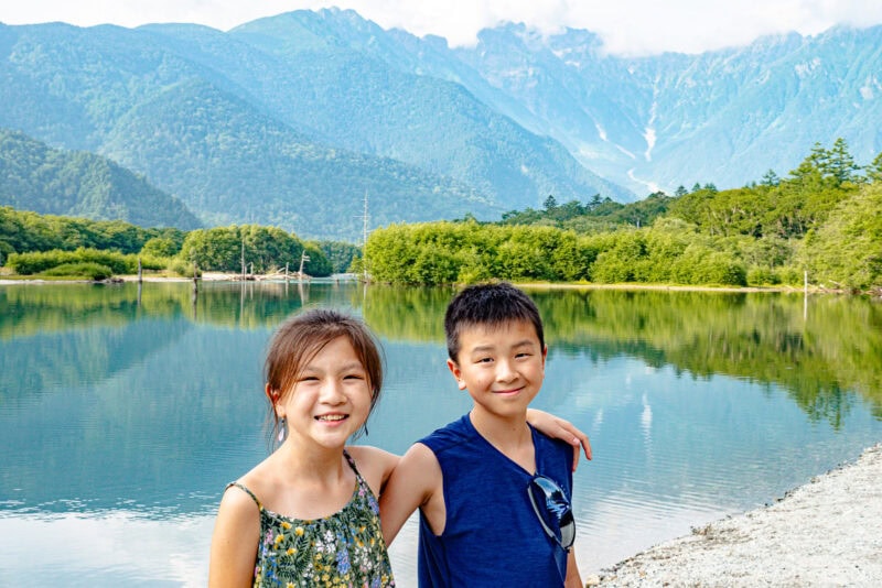 Two smiling children stand close together by the serene Taisho Pond, with lush green trees and tall mountains in the background under a partly cloudy sky.