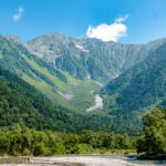 Kamikochi (上高地) – Exploring Japan’s Alpine Wonderland A lush, green mountain range under a bright blue sky with a few clouds, featuring dense forests and a rocky riverbed in the foreground—capturing the tranquil beauty of Kamikochi.