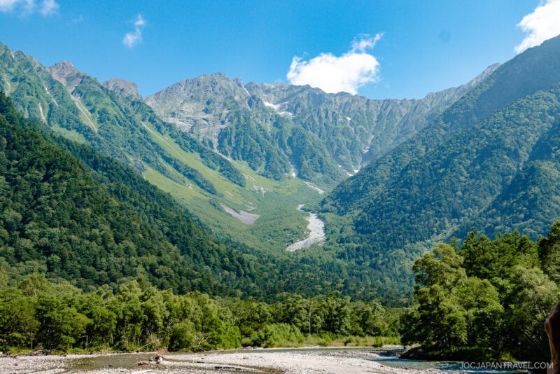 A lush, green mountain range under a bright blue sky with a few clouds, featuring dense forests and a rocky riverbed in the foreground&mdash;capturing the tranquil beauty of Kamikochi.