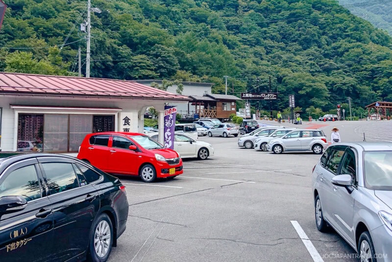 A parking lot with several cars parked, including a prominent red car, sits at the entrance to Kamikochi. Small buildings and signs in Japanese dot the area, with lush green forested hills rising in the background.