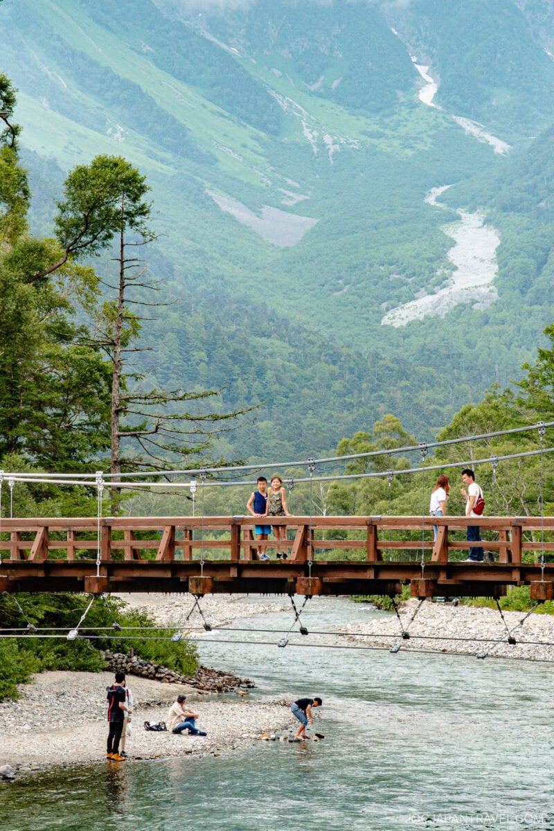 A wooden suspension bridge, known as Kappa Bridge, crosses a clear river with people standing and walking on it. Below, several people wade and sit on the rocky riverbank. Green mountains and trees fill the background.