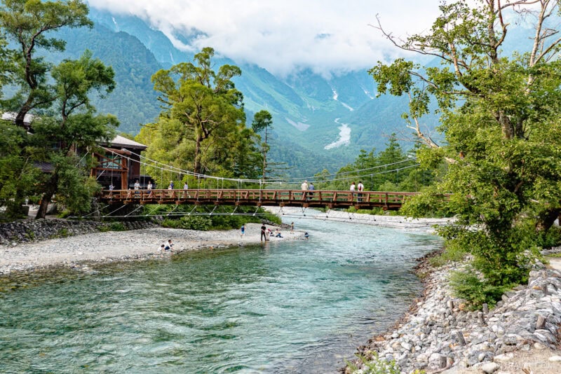 A wooden suspension bridge, known as Kappa Bridge, crosses a clear river with people walking on the bridge and along the rocky shore, surrounded by lush trees and mountains partially covered by clouds in the background.