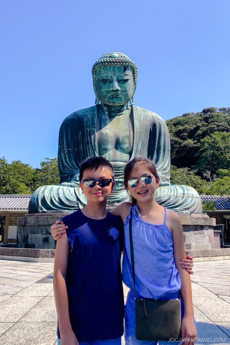 Two children wearing sunglasses and blue outfits pose together, smiling in front of the Great Buddha (Daibutsu) at Kotokuin in Kamakura, Japan, on a sunny day. Trees and blue sky are visible in the background.