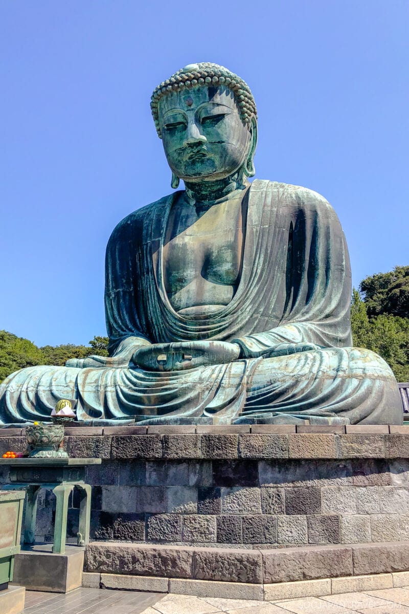 A large bronze statue of a seated Buddha in meditation pose sits outdoors on a stone pedestal at Kotokuin, with a clear blue sky and trees in the background.