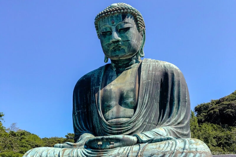 A large bronze statue of Buddha, known as the Great Buddha of Kamakura, sits outdoors at Kotokuin under a clear blue sky, surrounded by greenery. The Buddha is seated in a meditative pose with hands folded in its lap.