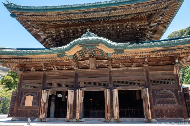 A large traditional Japanese wooden temple gate at Kenchoji Temple, featuring intricate carvings and a green-tiled roof, set against a clear blue sky.
