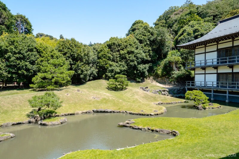 A traditional Japanese garden at Kenchoji Temple features a pond, manicured lawn, scattered rocks, small pine trees, and a wooden terrace building, all surrounded by lush green trees under a clear blue sky.