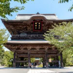 Kenchoji Temple (建長寺) – Kamakura’s Oldest Zen Temple A large traditional Japanese wooden gate at Kenchoji Temple, featuring intricate architectural details, stands surrounded by trees and greenery as people walk underneath on a sunny day.