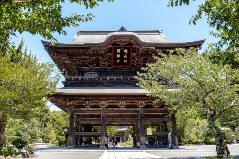 A large traditional Japanese wooden gate at Kenchoji Temple, featuring intricate architectural details, stands surrounded by trees and greenery as people walk underneath on a sunny day.