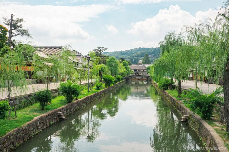 A peaceful canal lined with green trees and traditional Japanese buildings on both sides&mdash;discover this tranquil scene in our Kurashiki travel guide, featuring a small stone bridge beneath a partly cloudy sky.